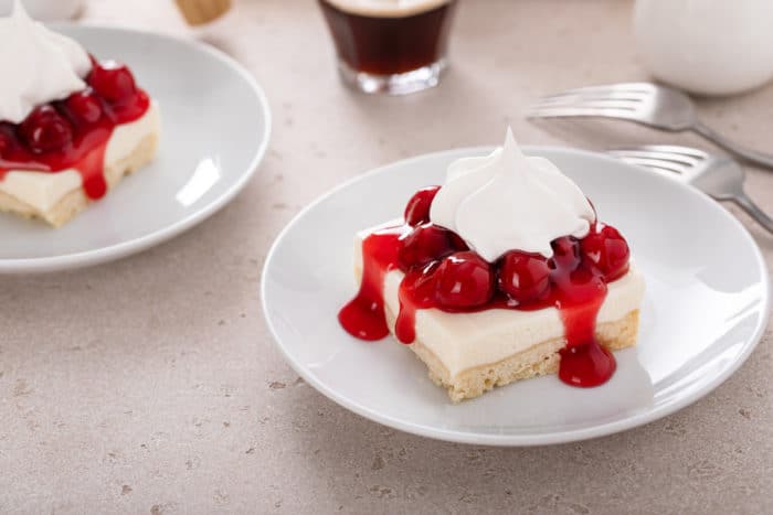 Two white plates, each with a slice of nana's easy cheesecake topped with cherry pie filling and a dollop of whipped topping.