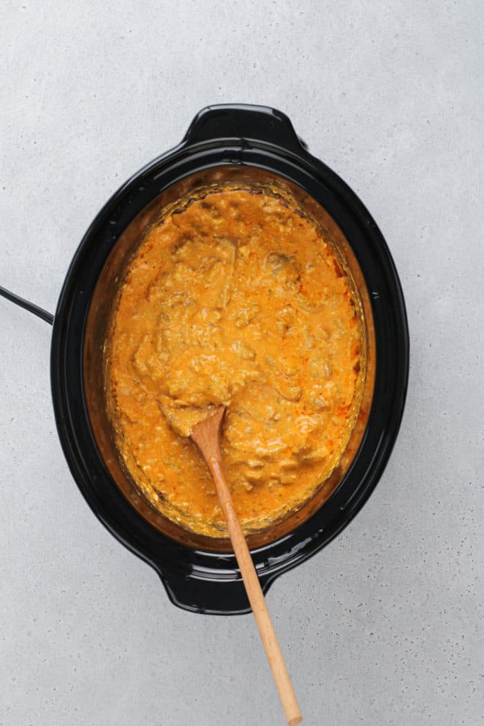 Wooden spoon stirring buffalo chicken dip cooking in a slow cooker.