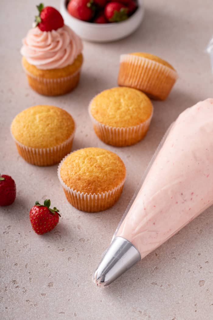Piping bag filled with strawberry frosting on a countertop next to vanilla cupcakes.