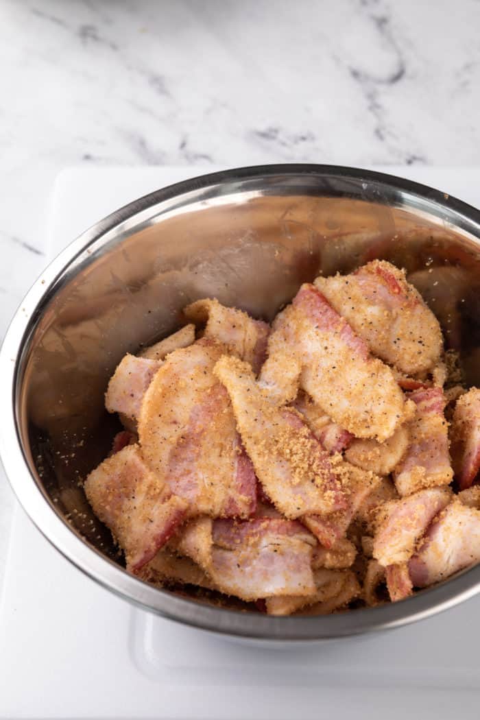 Pieces of bacon tossed with brown sugar and spices in a metal mixing bowl.