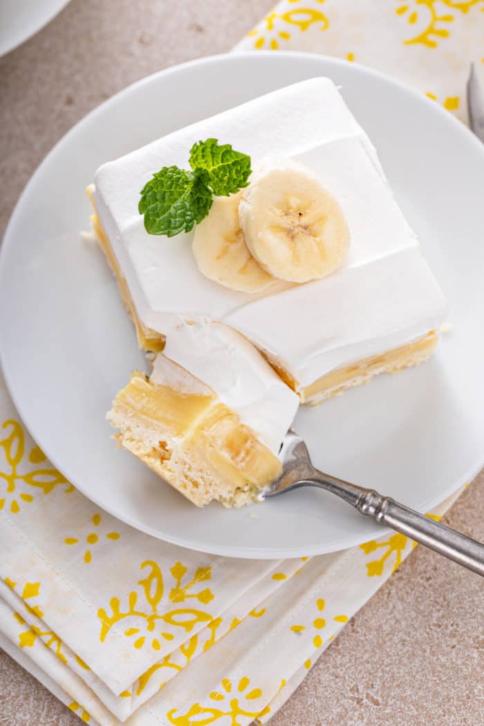 Fork cutting a bite off of a banana cream bar on a white plate.