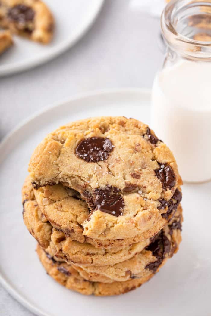 Bite taken from the top cookie in a stack of chocolate chip cookies with brown butter and toffee. A glass of milk is next to the cookies.