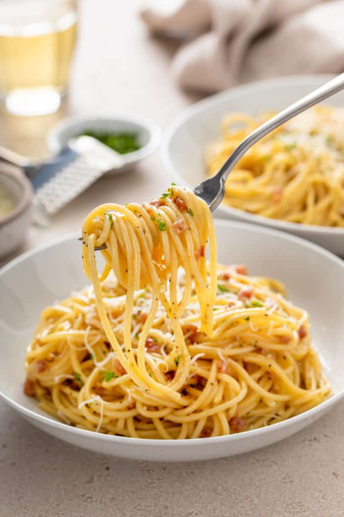 Bite of pasta carbonara twirled onto a fork and being held above a bowl of the pasta.