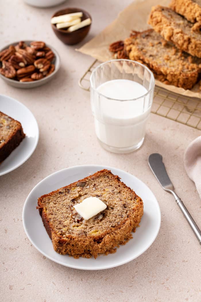 Slice of banana nut bread topped with butter on a white plate. A glass of milk and more slices of bread are visible in the background.