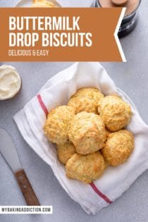Overhead view of a tea-towel-lined basket filled with buttermilk drop biscuits on a gray countertop. Text overlay includes recipe name.