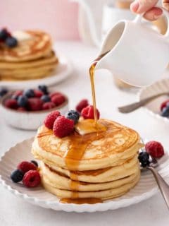 Syrup being poured over a stack of buttermilk pancakes on a white plate.