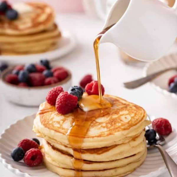 Syrup being poured over a stack of buttermilk pancakes on a white plate.