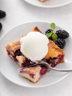 Fork cutting a bite from the corner of a piece of blackberry cobbler topped with ice cream.