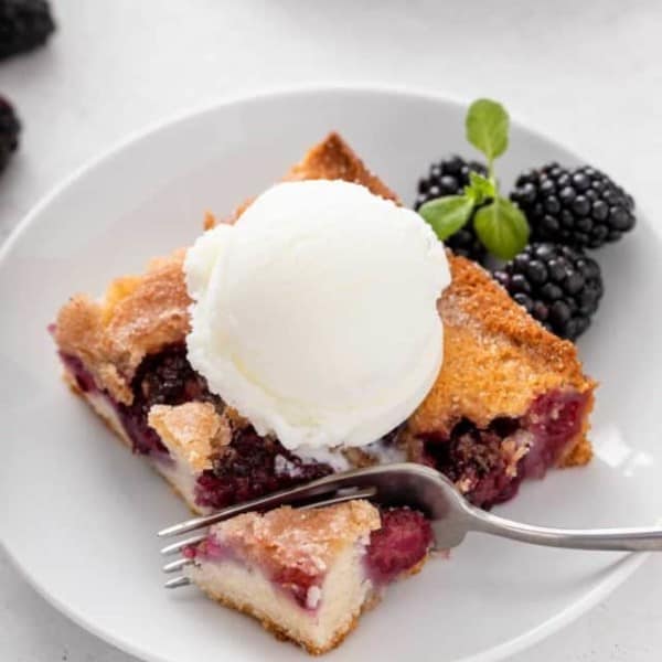 Fork cutting a bite from the corner of a piece of blackberry cobbler topped with ice cream.