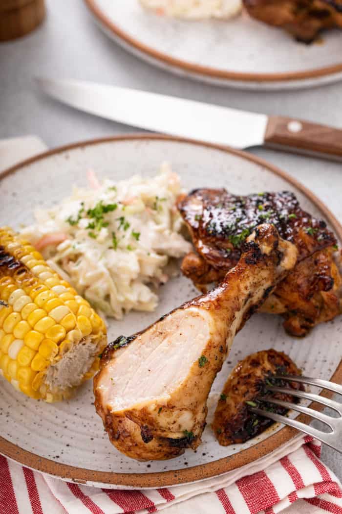 Bite cut from a grilled chicken drumstick on a stoneware plate next to corn and slaw.