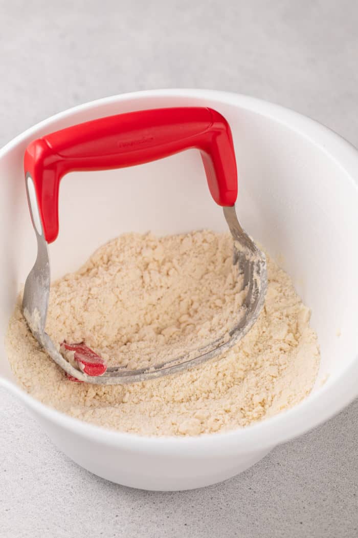 Butter cut into dry ingredients with a pastry blender in a white mixing bowl.
