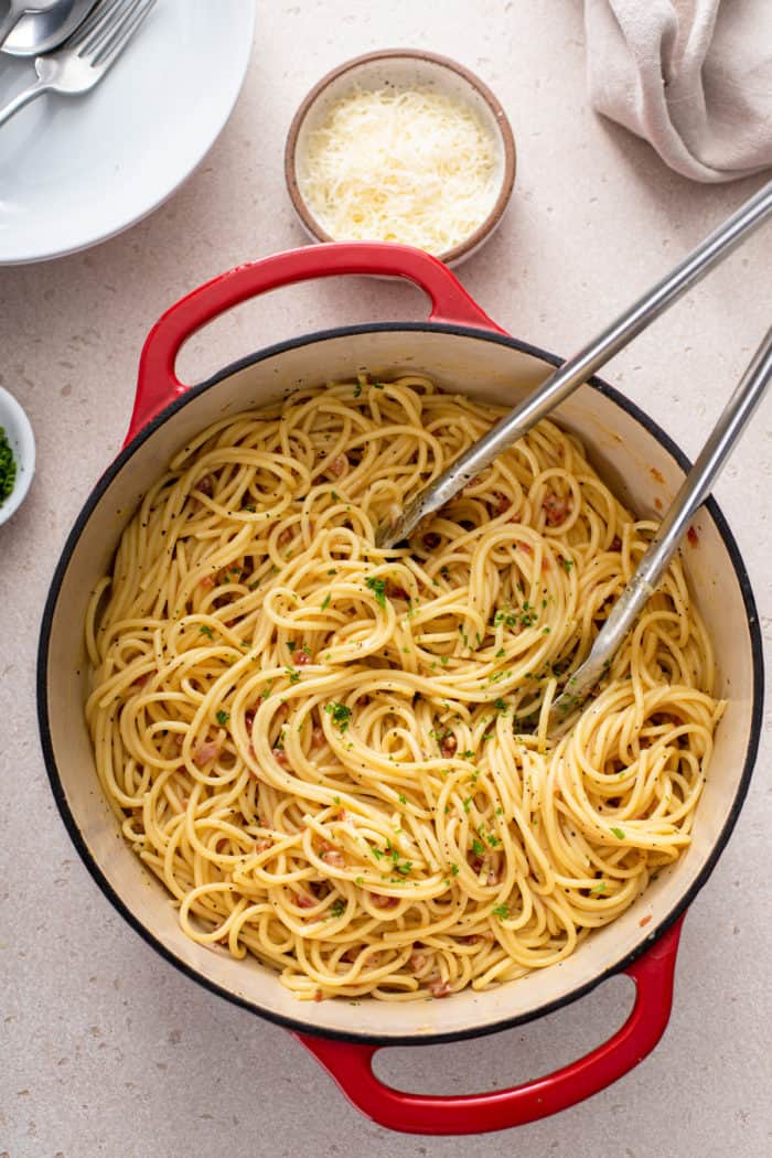 Overhead view of tongs stirring a pot of pasta carbonara in a red dutch oven.