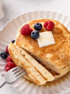 Fork with a bite of buttermilk pancakes next to the stack of pancakes on a plate.