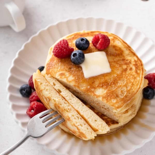 Fork with a bite of buttermilk pancakes next to the stack of pancakes on a plate.