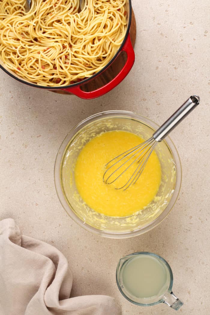 Pasta water being whisked into a bowl of eggs and cheese for pasta carbonara, set next to a pot of pasta tossed with cooked pancetta.