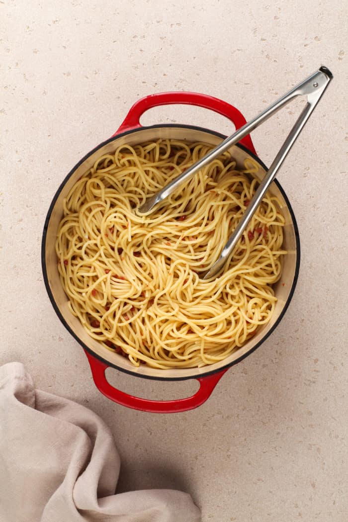 Tongs tossing cooked spaghetti with crispy pancetta in a red dutch oven.