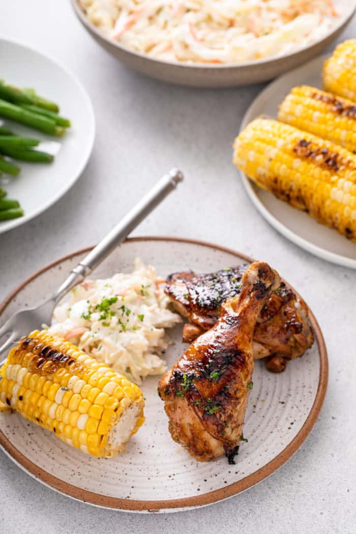 A grilled chicken drumstick and thigh arranged on a plate next to grilled corn and coleslaw.