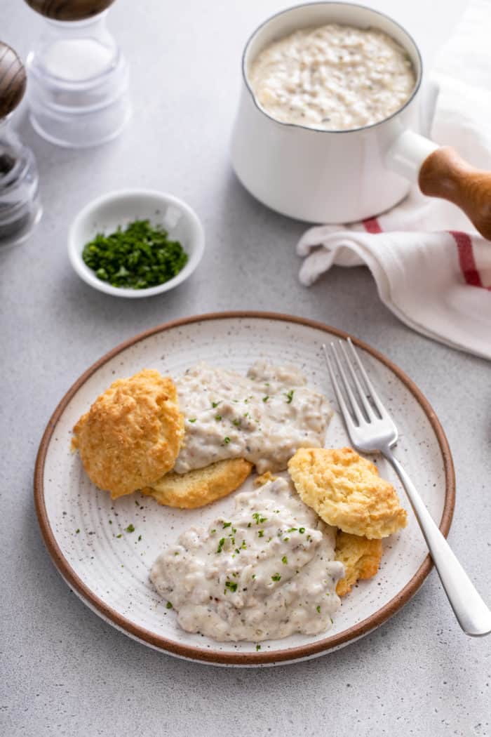 Two biscuits topped with sausage gravy on a stoneware plate. A small pot of gravy is visible in the background.