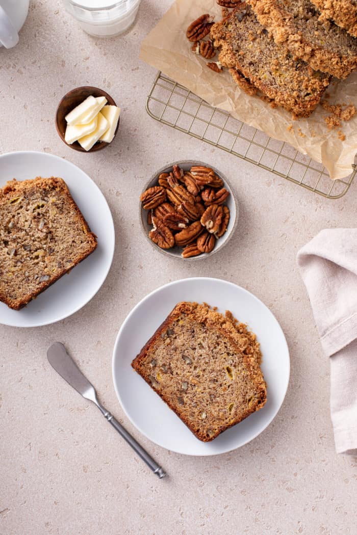 Two slices of banana nut bread on white plates next to the sliced loaf of bread.