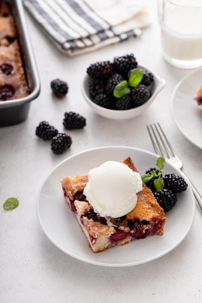 Slice of easy blackberry cobbler topped with a scoop of vanilla ice cream on a white plate. A bowl of blackberries is in the background.