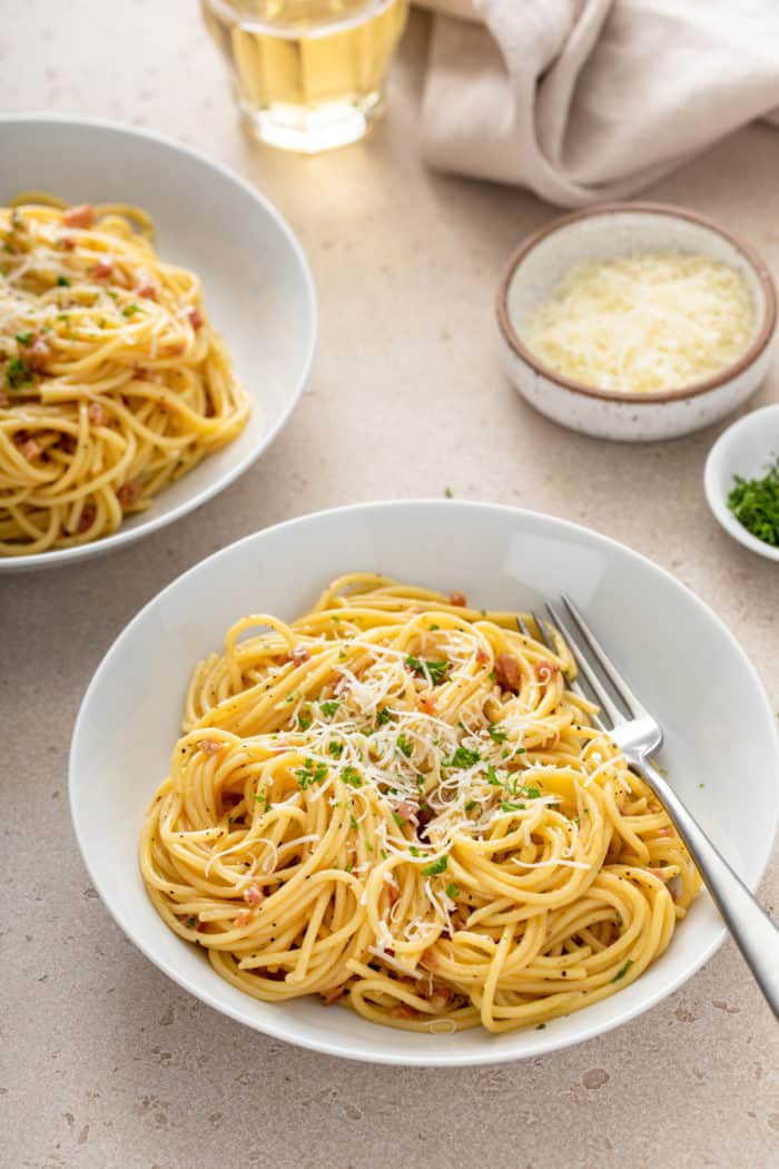 White bowl filled with pasta carbonara. Another bowl of the pasta and a small bowl of parmesan cheese are both visible in the background.