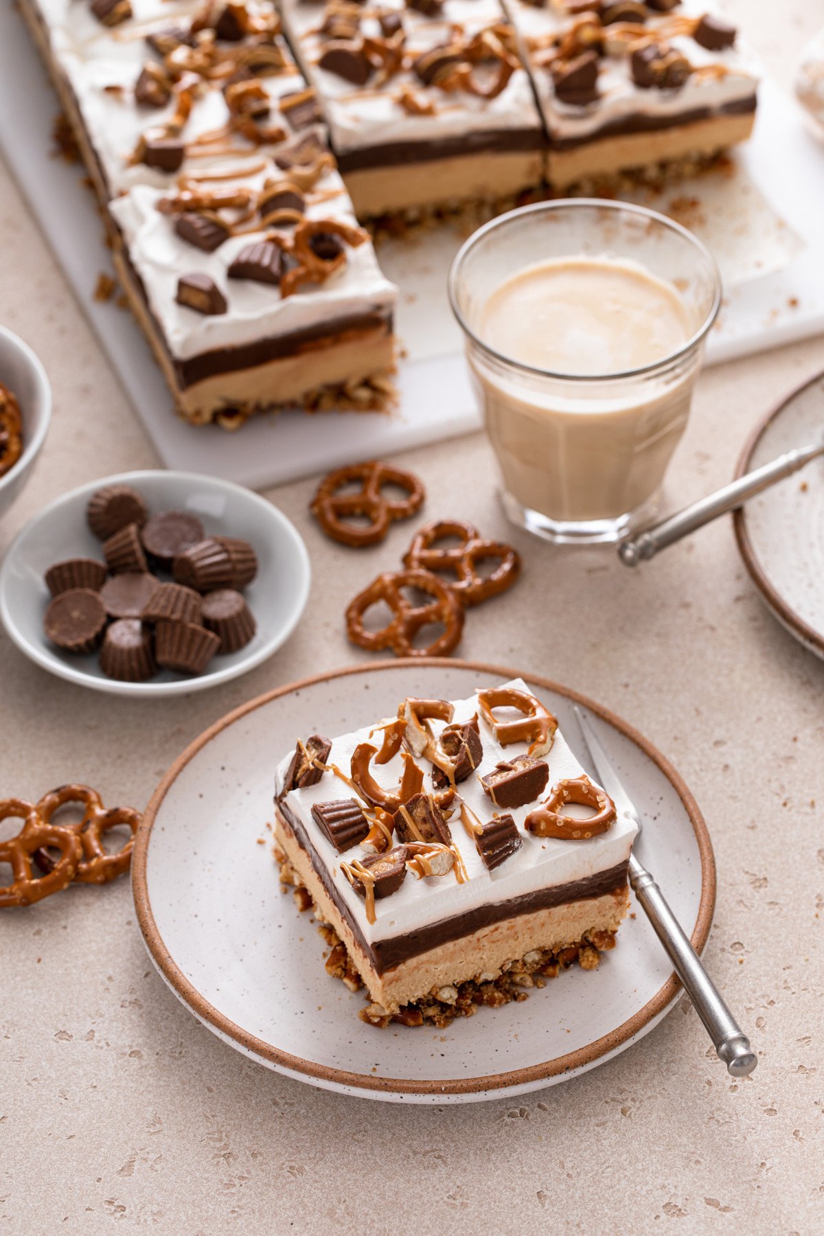 Plated slice of peanut butter pretzel dessert with a cup of coffee and more of the dessert in the background.