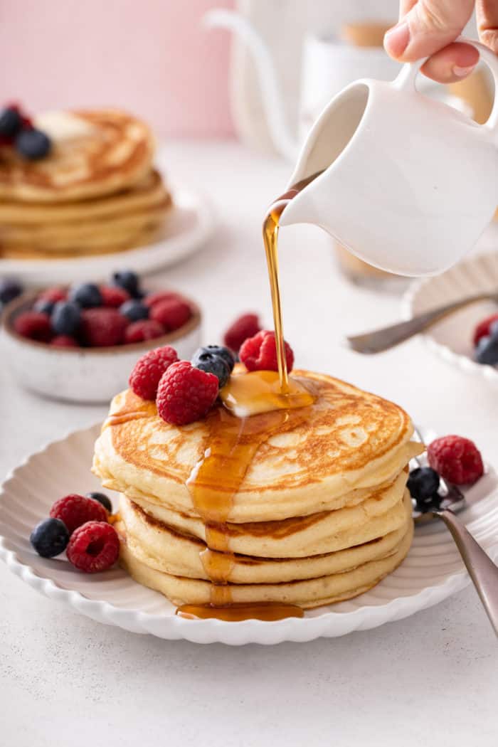 Syrup being poured over a stack of buttermilk pancakes on a white plate.