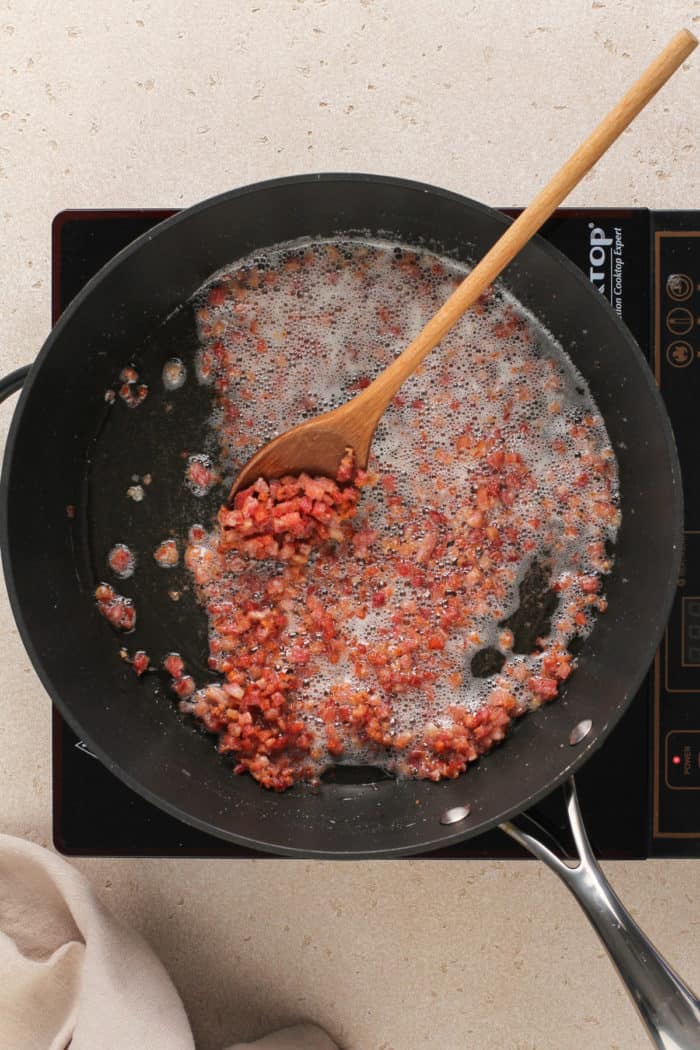 Pancetta being rendered in a black skillet on an induction burner.