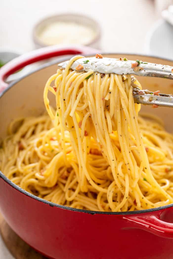 Tongs lifting a serving of pasta carbonara out of a red dutch oven filled with the pasta.