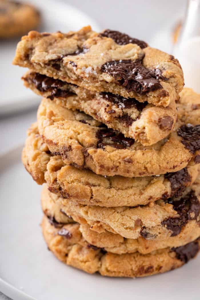 Stack of chocolate chip cookies with brown butter and toffee. the top cookie is broken in half, showing the melty chocolate chips and gooey inner texture.