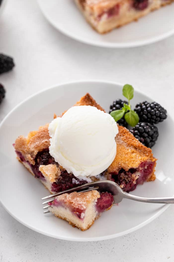 Fork cutting a bite from the corner of a piece of blackberry cobbler topped with ice cream.