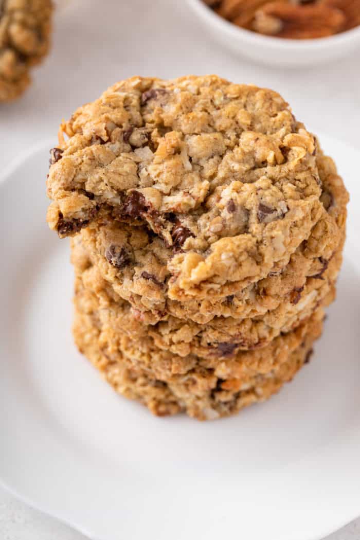 Stack of cowboy cookies on a white plate with a bite taken from the top cookie.