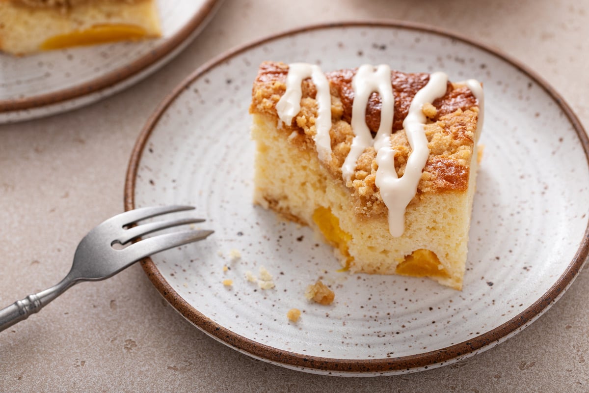 Slice of easy peach coffee cake on a speckled plate. A bite has been taken from the corner of the cake and a fork is resting on the edge of the plate.