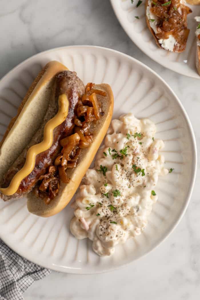 Brat on a bun topped with mustard and caramelized onions, set on a plate next to potato salad.