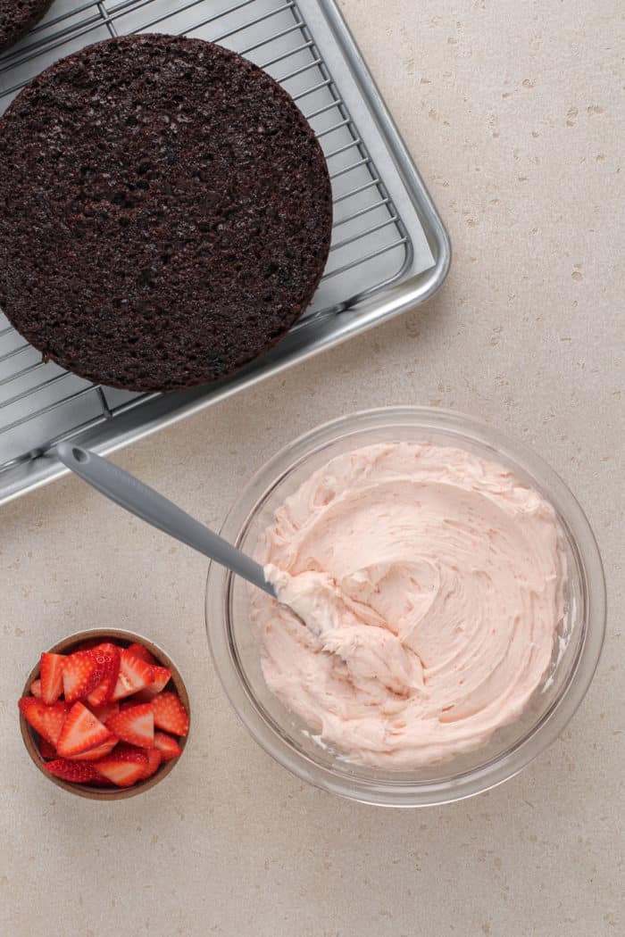 Chocolate cake layers on a wire cooling rack next to a bowl of strawberry frosting.