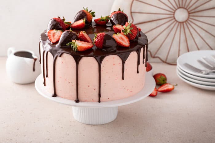 White cake stand holding up a chocolate cake covered in strawberry frosting and topped with chocolate ganache and fresh strawberries.