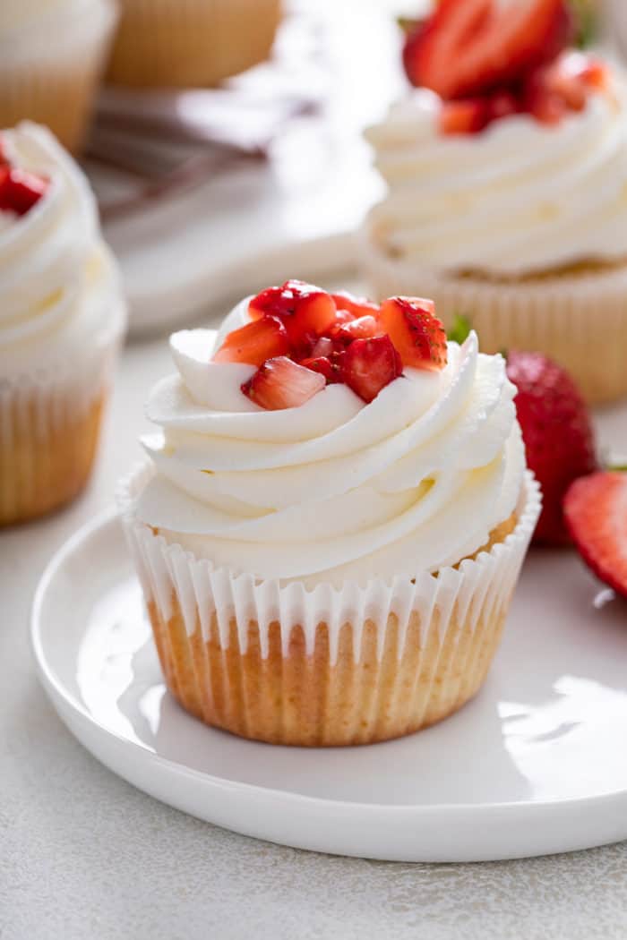 Strawberry shortcake cupcake topped with whipped cream frosting and diced strawberries set on a white plate. 