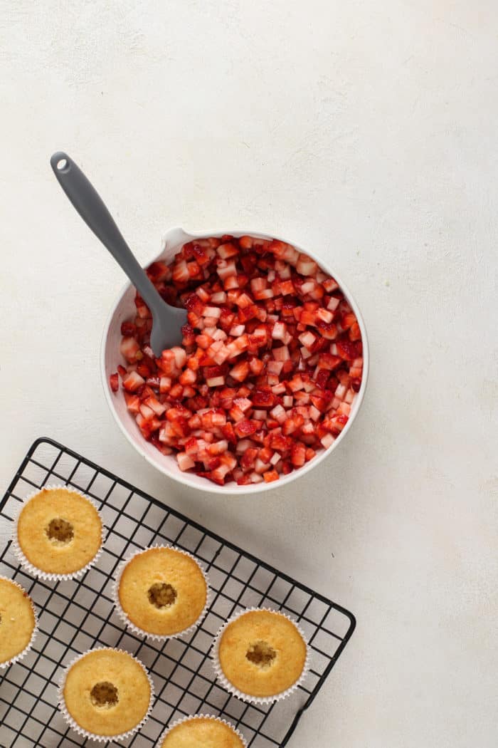 Cored shortcake cupcakes on a wire rack next to a bowl of diced macerated strawberries.