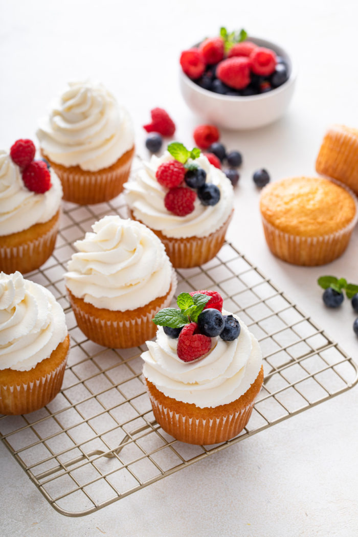Vanilla cupcakes topped with whipped cream frosting and fresh berries scattered on a wire cooling rack.