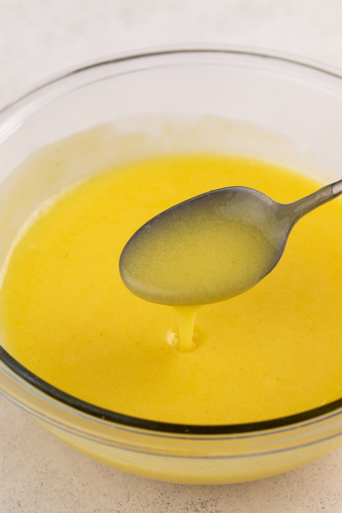 Spoon pouring freshly cooked lime curd into a glass bowl of the curd to show the texture.