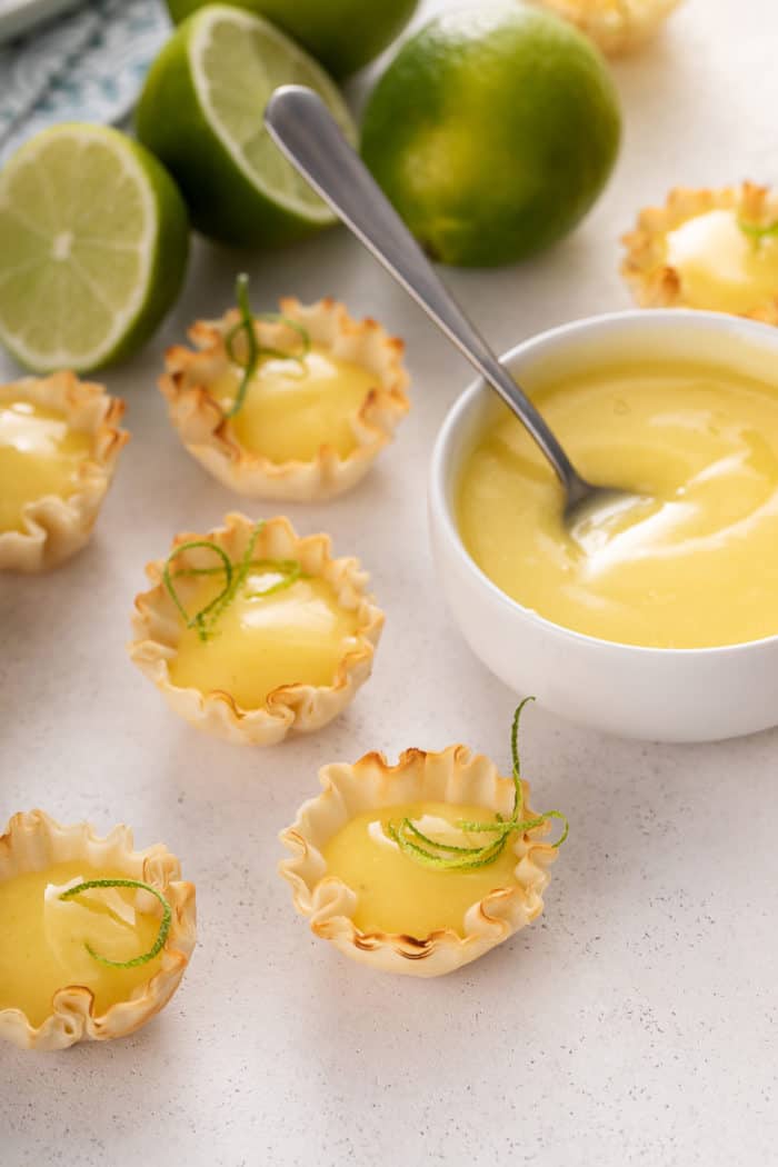 Phyllo shells filled with lime curd arranged next to a white bowl filled with lime curd.