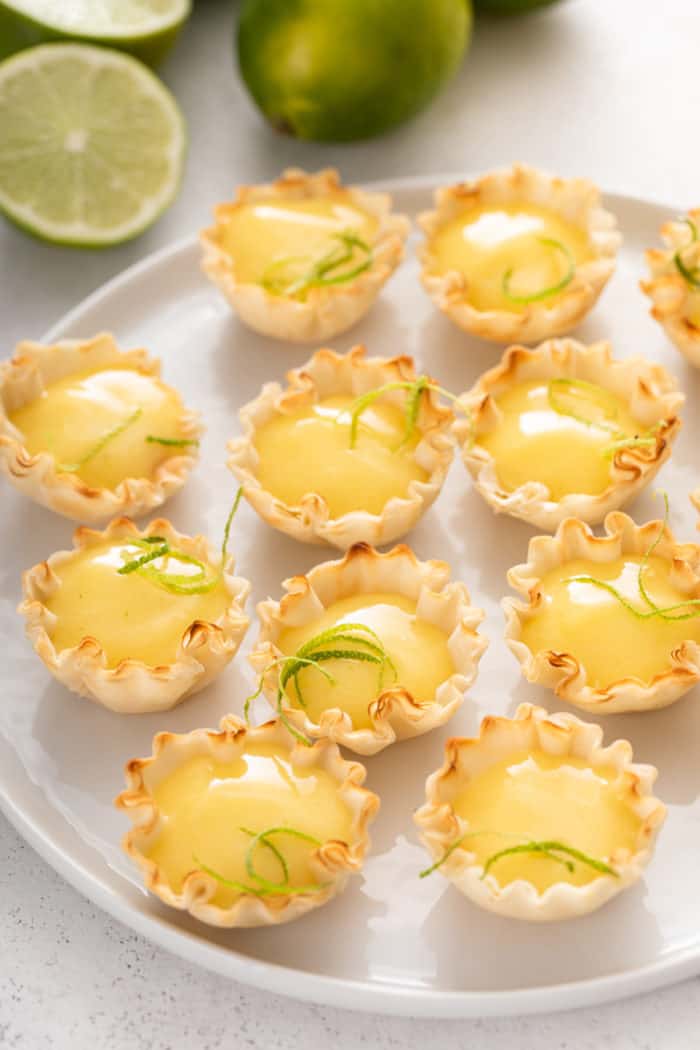 White platter filled with mini phyllo tart shells that are filled with lime curd and topped with lime zest.