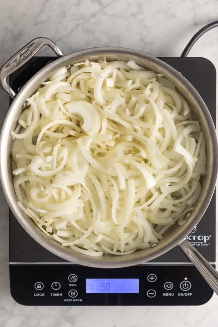 Sliced onions starting to soften in a large skillet set on an induction burner.