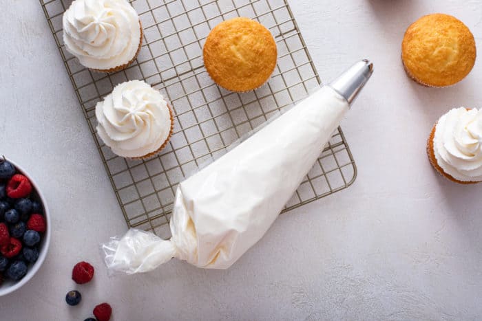 Piping bag filled with whipped cream frosting set onto a wire rack alongside vanilla cupcakes.