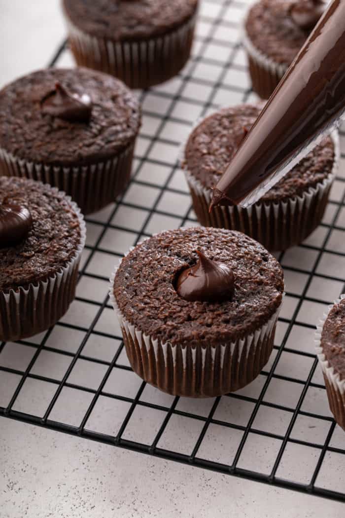 Chocolate ganache being piped into chocolate cupcakes on a wire rack.