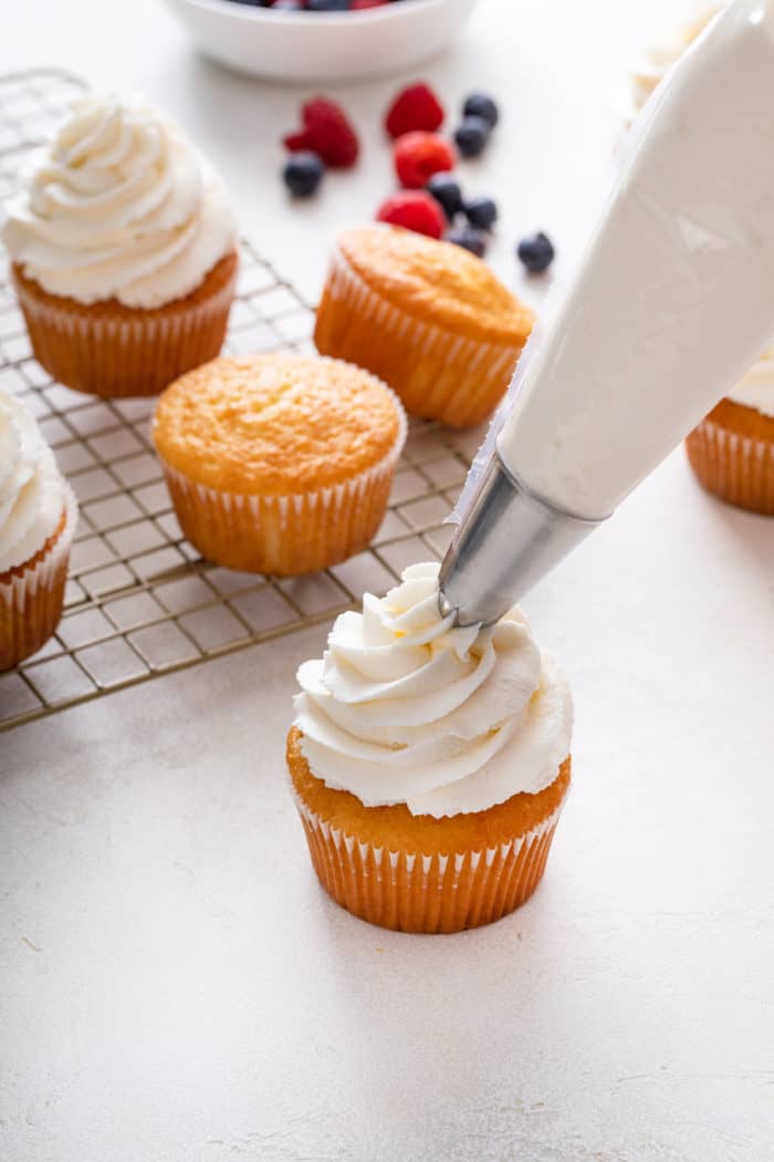 Whipped cream frosting being piped onto a vanilla cupcake with a piping bag.
