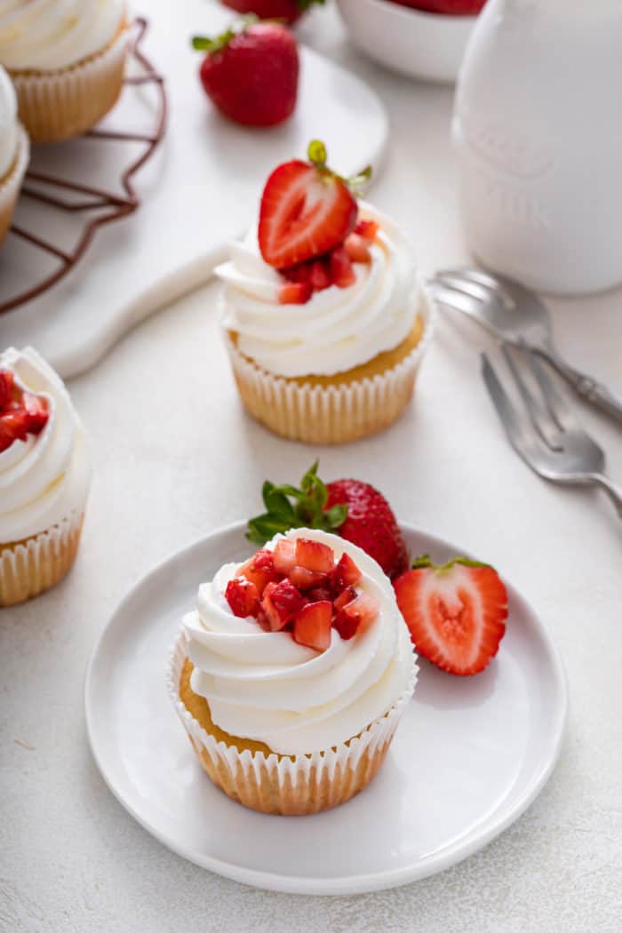 White plate holding a strawberry shortcake cupcake and fresh strawberries, with additional cupcakes in the background.