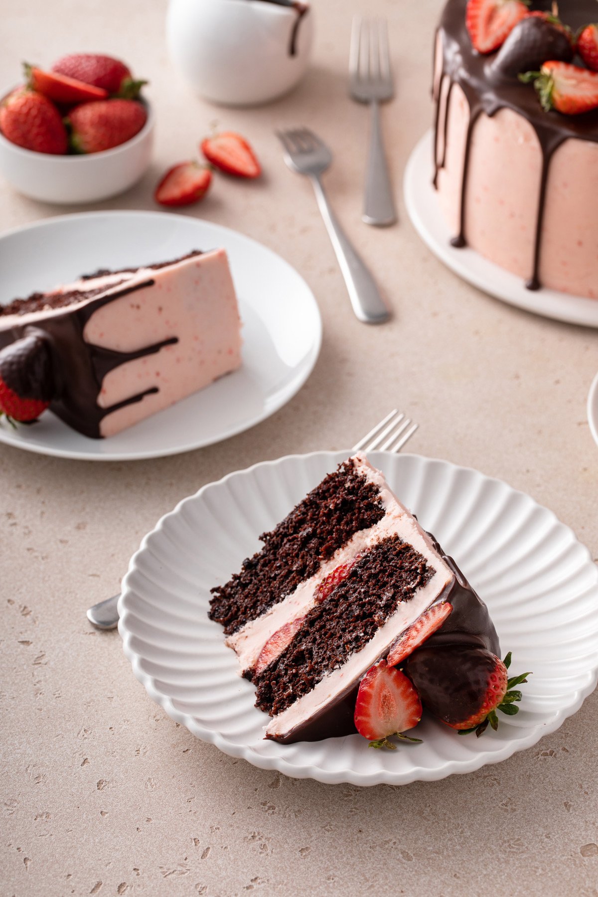 Two white plates, each holding a slice of chocolate strawberry cake, with the rest of the cake visible in the background.