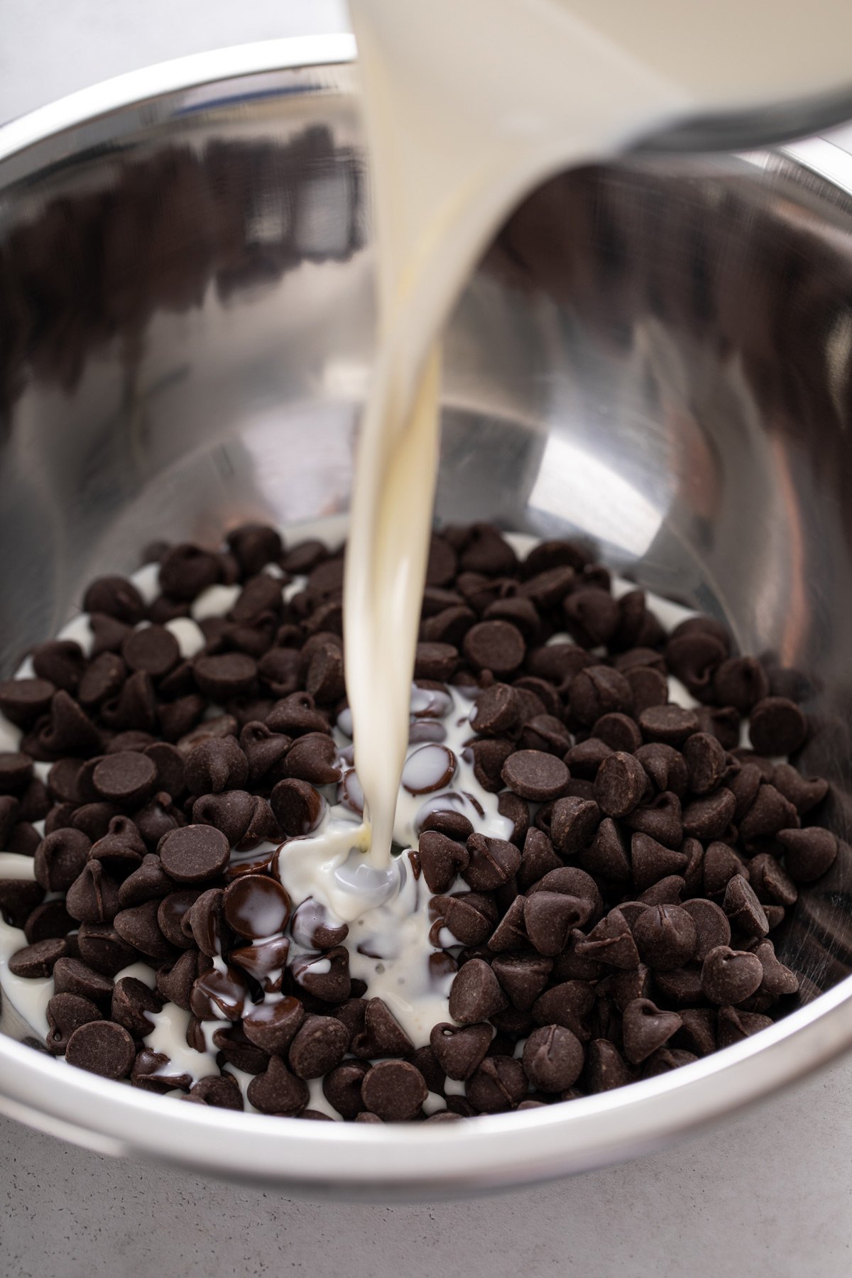 Hot cream being poured over chocolate chips in a metal bowl.