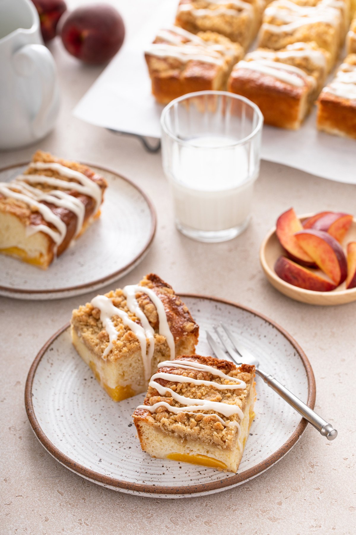 Two plates, each holding slices of peach coffee cake, set on a countertop with a glass of milk and more cake in the background.